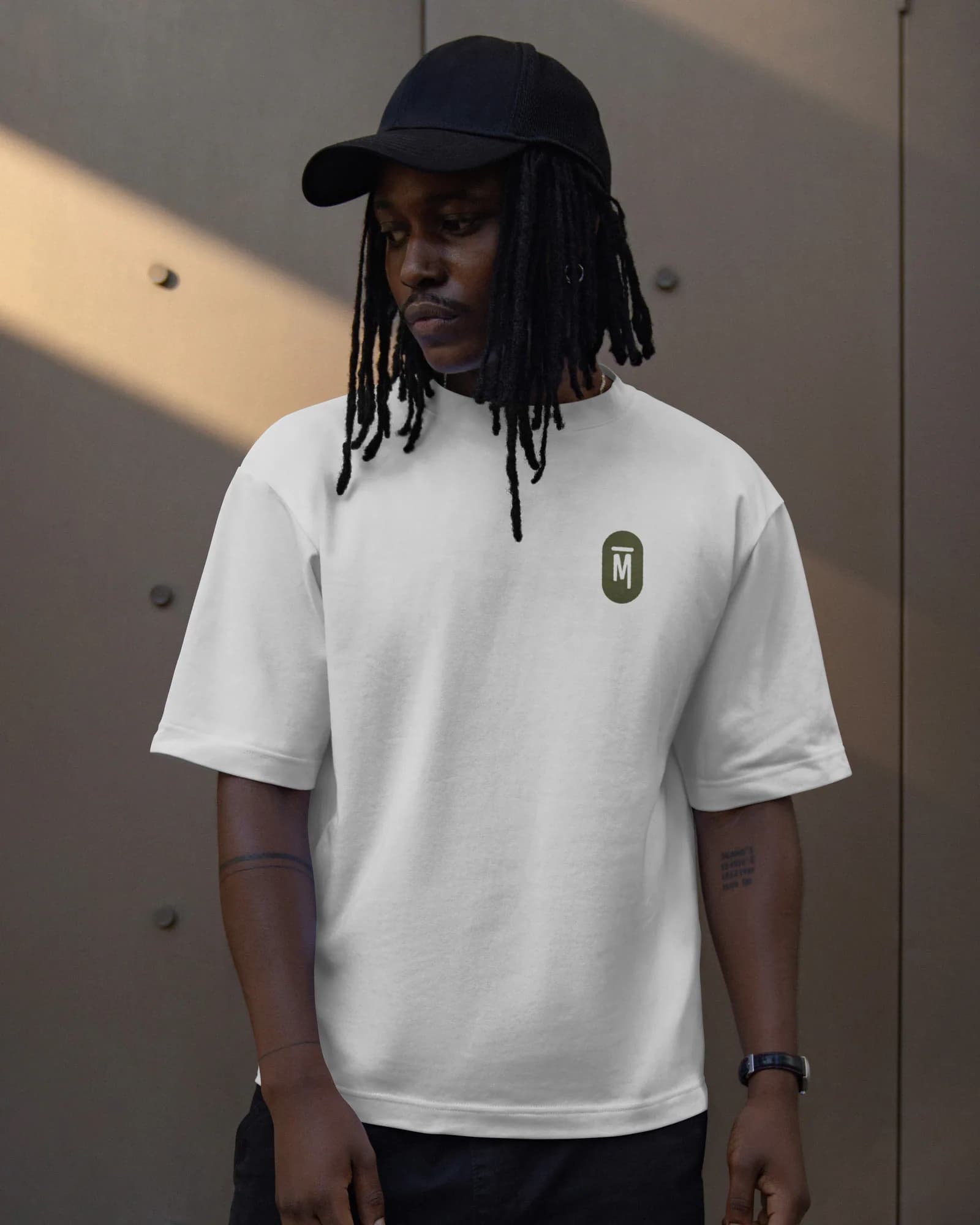 Modèle en casquette noire portant un t-shirt blanc ample, avec le petit monogramme La PAMPA vert olive sur la poitrine, devant un mur de métal