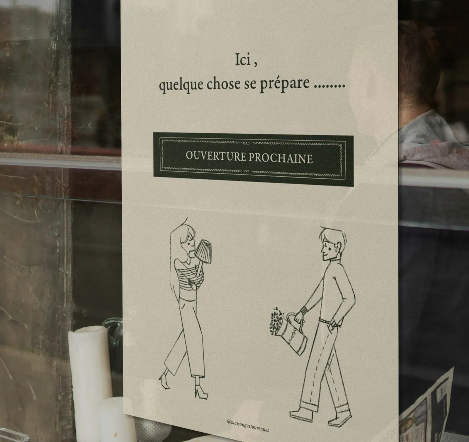 Affiche beige collée dans une vitrine : Ici, quelque chose se prépare, un cartouche vert Ouverture prochaine et les deux personnages au trait.
