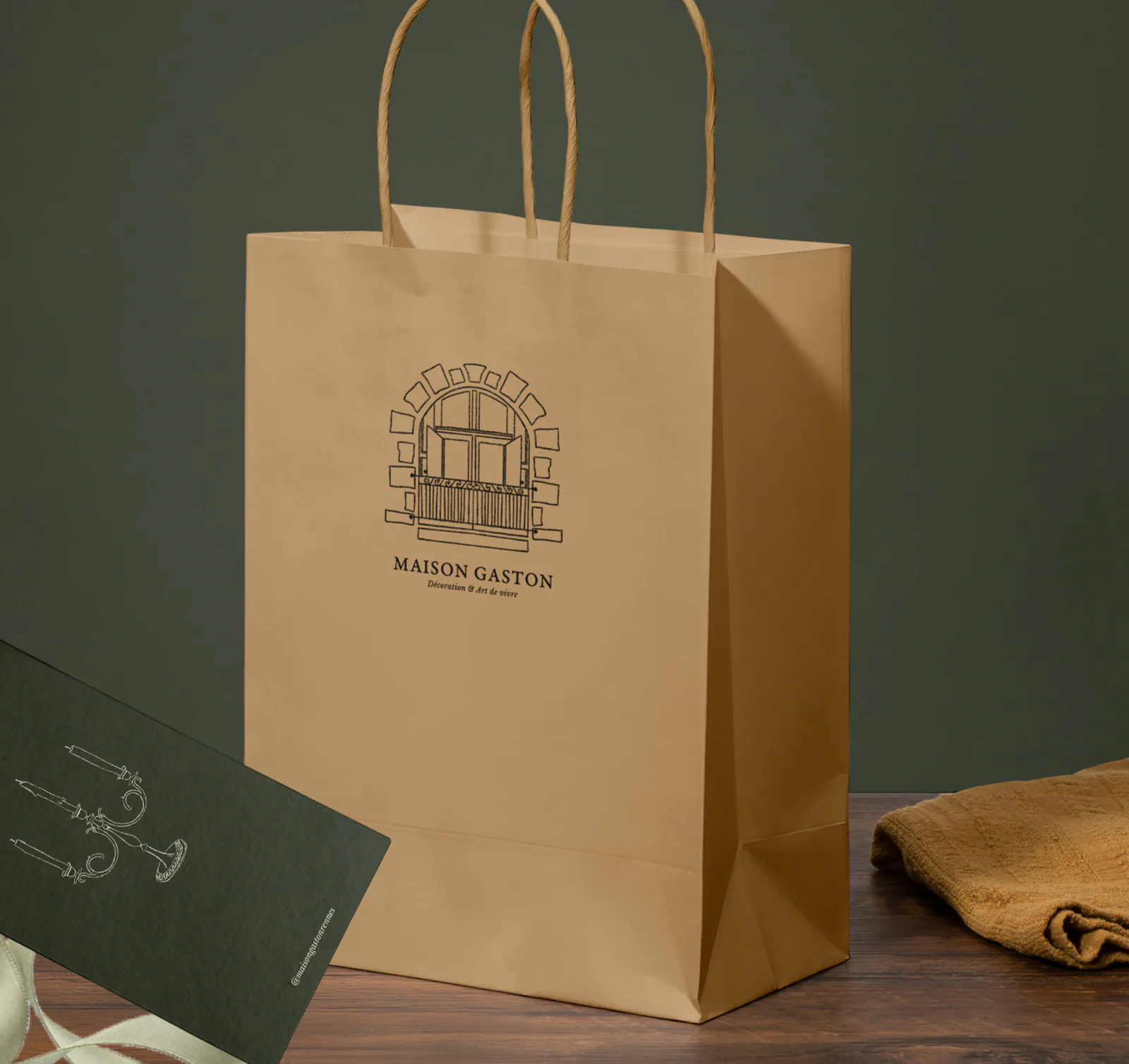 Sac kraft imprimé du logo Maison Gaston en linogravure, avec une étiquette vert olive illustrée nouée à un ruban sauge.