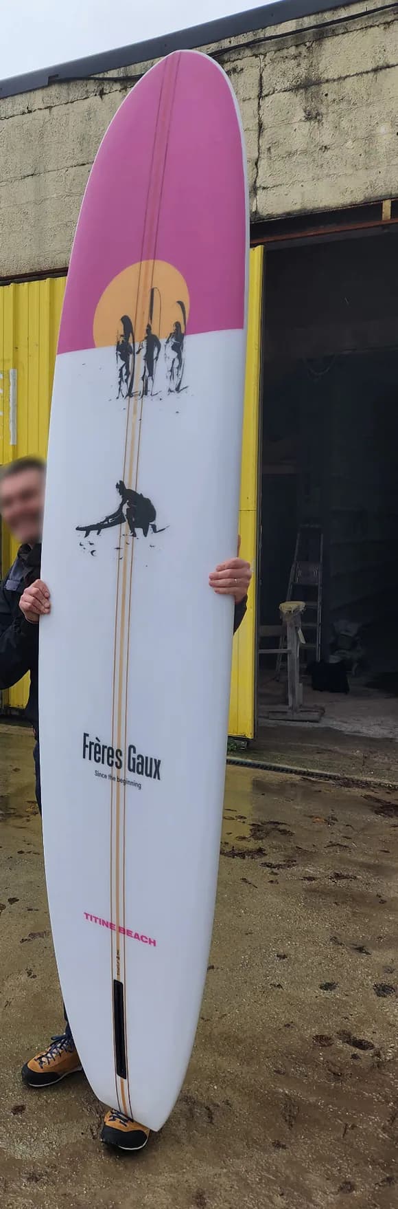 La planche Frères Gaux terminée, plus haute que la personne au visage flouté qui la tient debout devant un atelier au bardage jaune