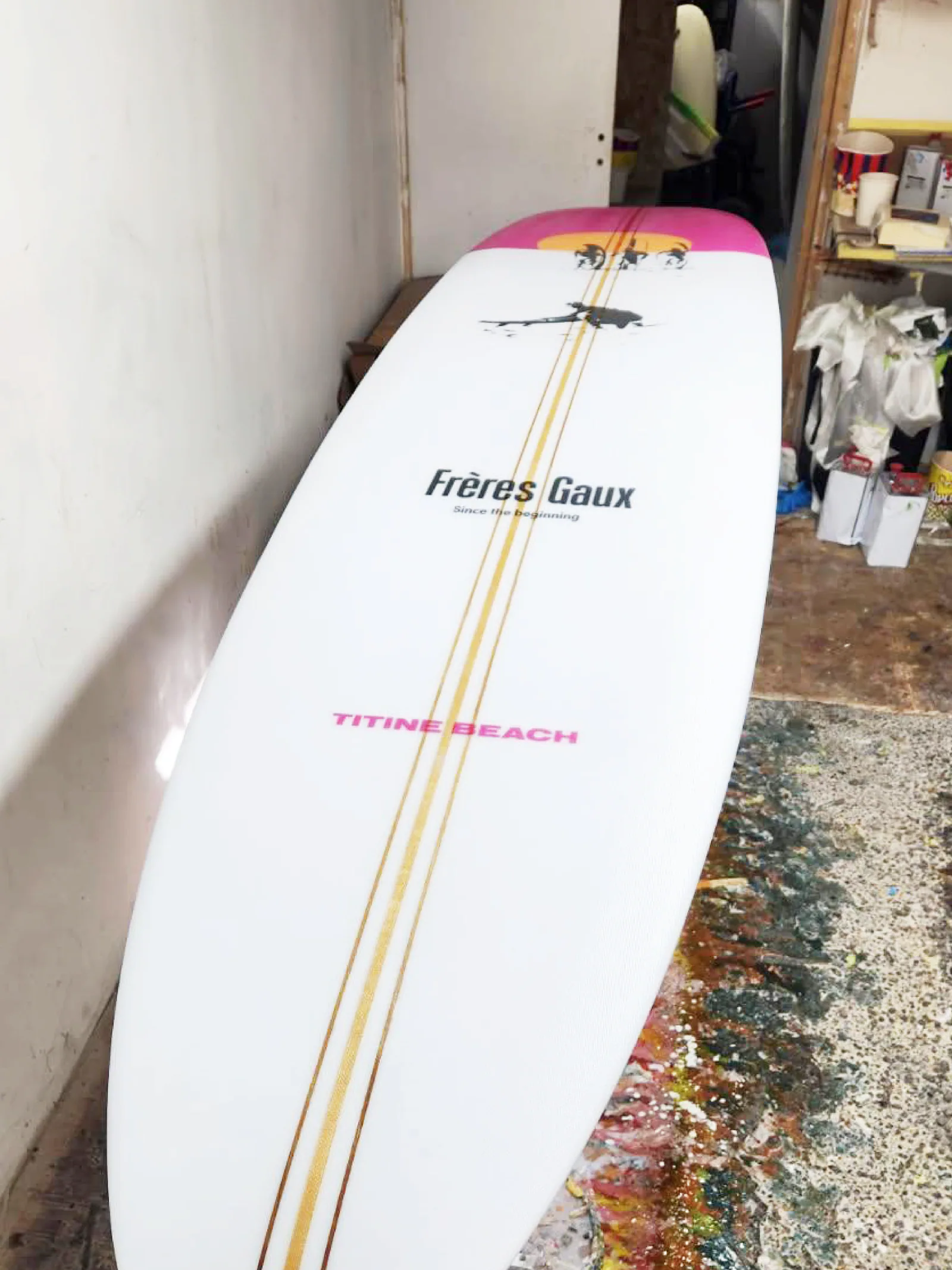 Planche de surf Frères Gaux posée en atelier : nez rose, soleil jaune, silhouettes noires, logo Frères Gaux et inscription rose Titine Beach