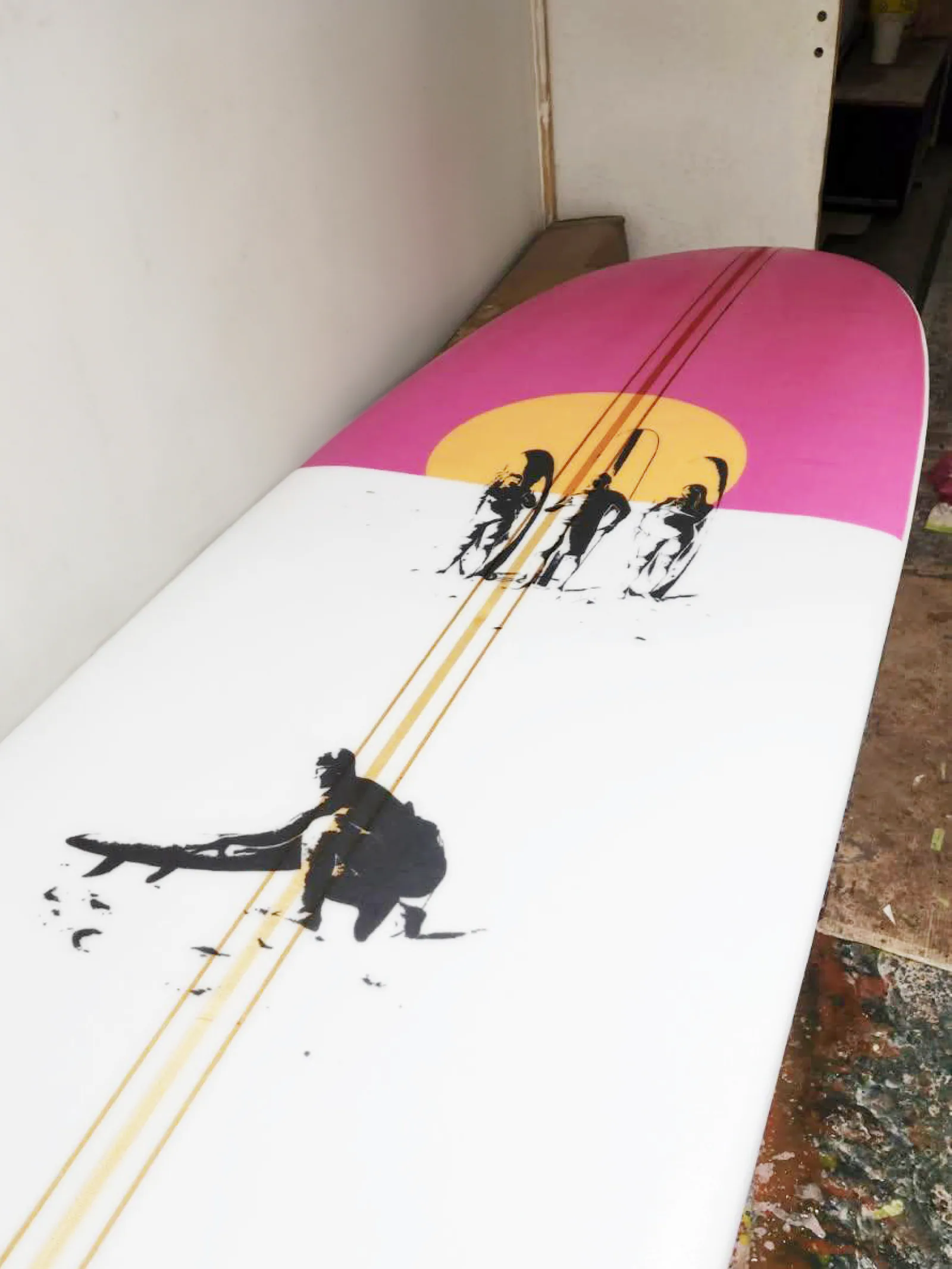 Nez rose de la planche Frères Gaux avec un soleil jaune orangé, trois silhouettes de surfeurs et un surfeur accroupi près de sa planche